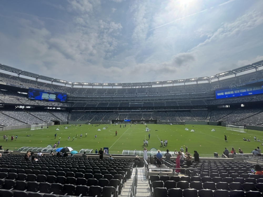 a wide angle image of metlife stadium from the stands showing youth dotting every corner of the soccer field duing a sunny day